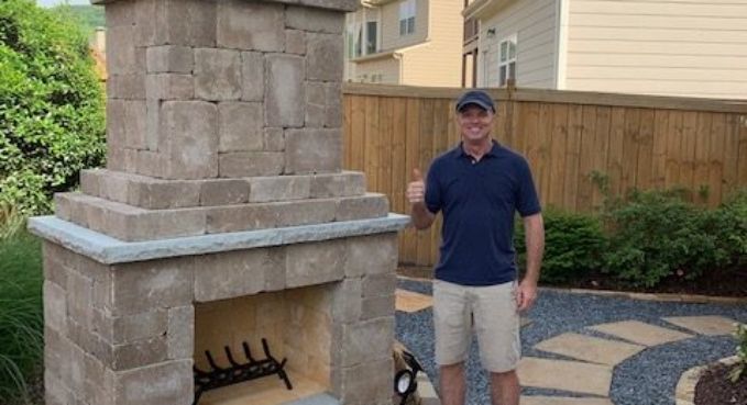 Man standing beside a stone outdoor fireplace in a landscaped backyard patio