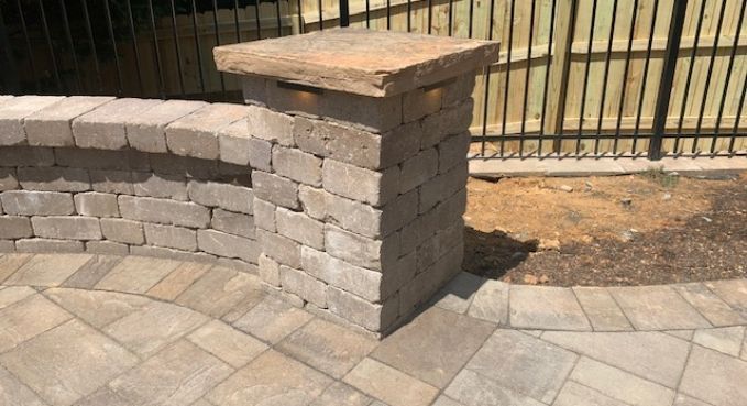 Stone patio wall with a square column and wood-topped cap beside a paved walkway and metal fence.