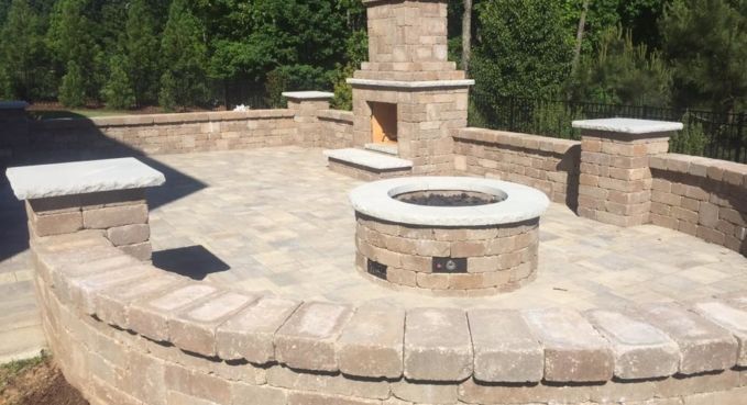 Stone outdoor fire pit and seating area with built-in benches on a patio, surrounded by greenery