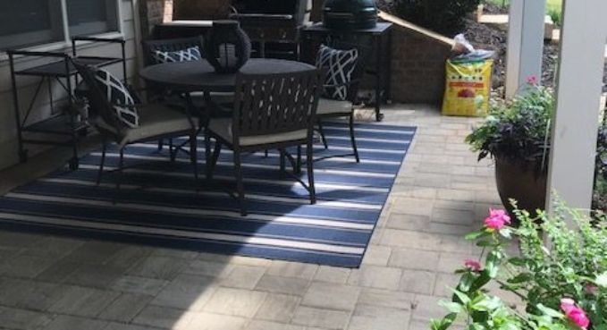 Patio with black metal table and chairs on striped outdoor rug beside potted flowers and stone pavers