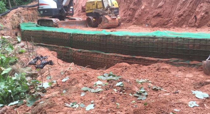 Excavator working beside a reinforced trench with green mesh and exposed red soil on a construction site