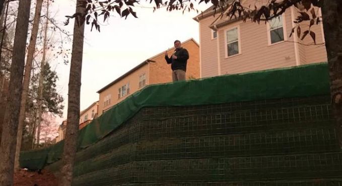 Person standing on a grassy hillside beside a beige house, near a retaining wall and trees