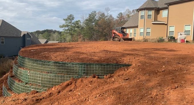 Retaining wall under construction on a dirt slope with an excavator and houses in the background