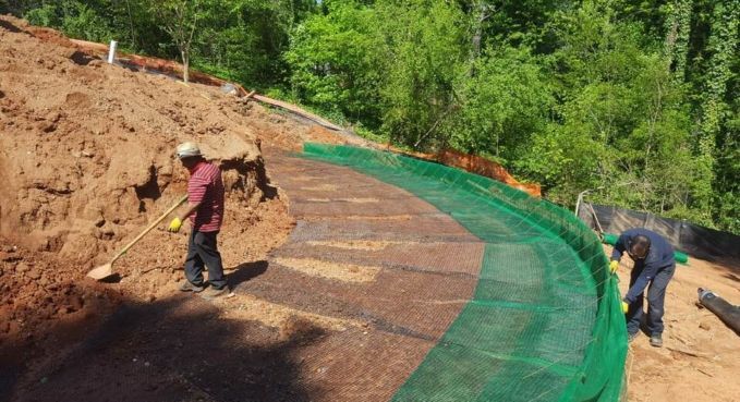 Workers install green mesh on a curved dirt slope beside a wooded hillside.
