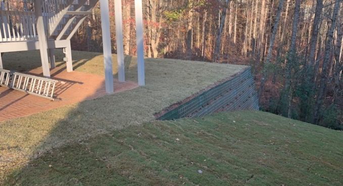 Grassy yard with a brick retaining wall, patio, and white support posts beside a wooded slope