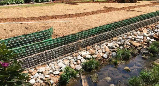Green erosion-control netting along a rocky stream beside a dirt embankment and grassy field