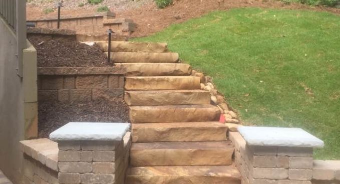 Stone staircase with wooden steps leading up a landscaped hillside, bordered by concrete pillars and grass