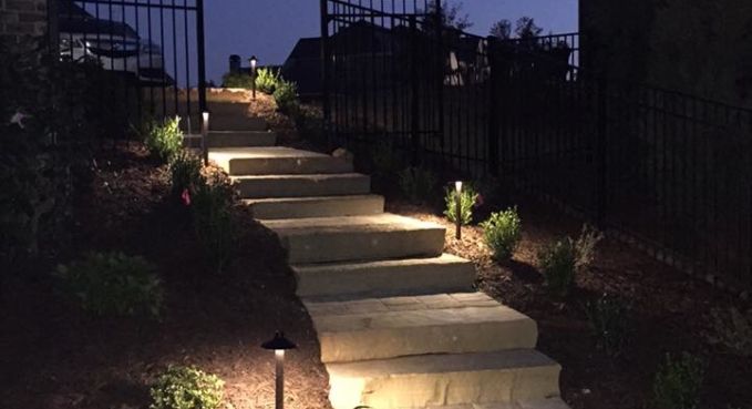 Illuminated stone steps leading uphill through a garden at dusk, bordered by dark iron railings.