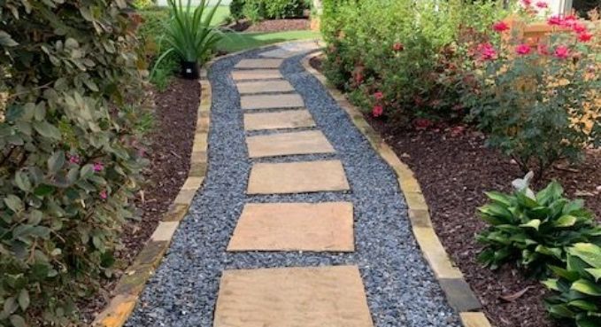 Curved garden path with tan stepping stones set in dark gravel, bordered by lush green plants and pink flowers