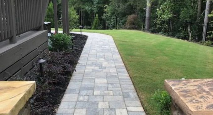 Stone walkway beside a house, bordered by mulch and lawn with trees in the background.