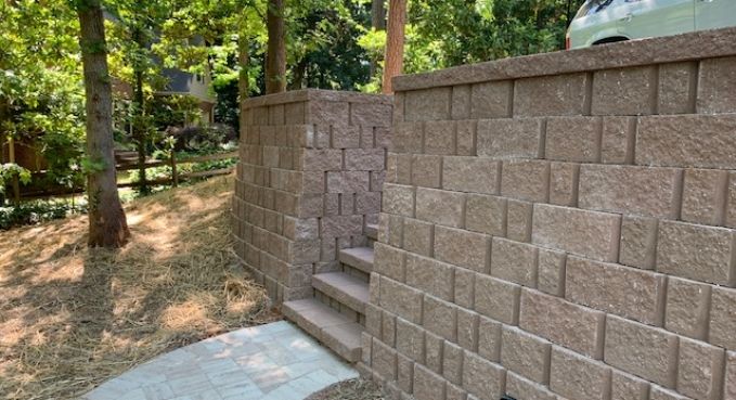 Stone retaining wall with steps leading up a wooded hillside path, beside a concrete walkway.