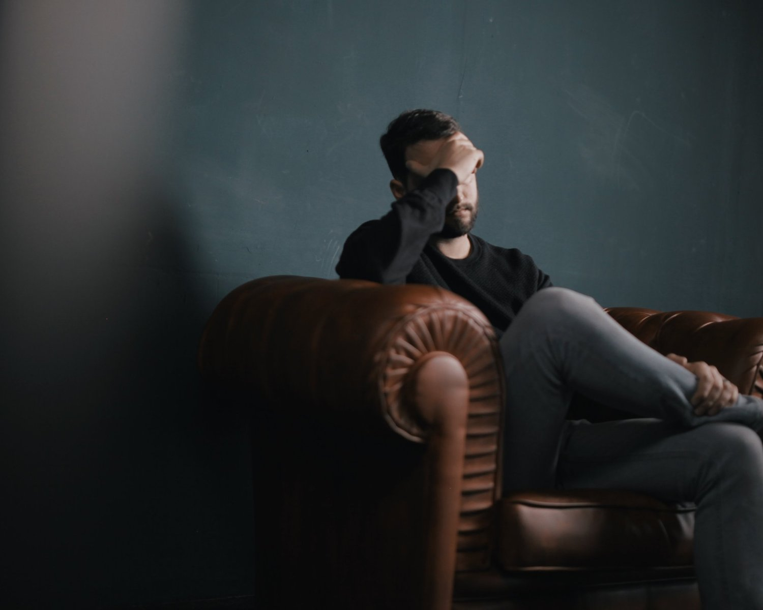 Man in dark clothes sits on a brown leather couch, hand on forehead, in a dim room with a dark wall.