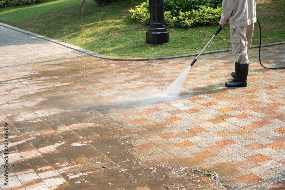 A worker in boots uses a pressure washer to clean dirt from a paved garden walkway.