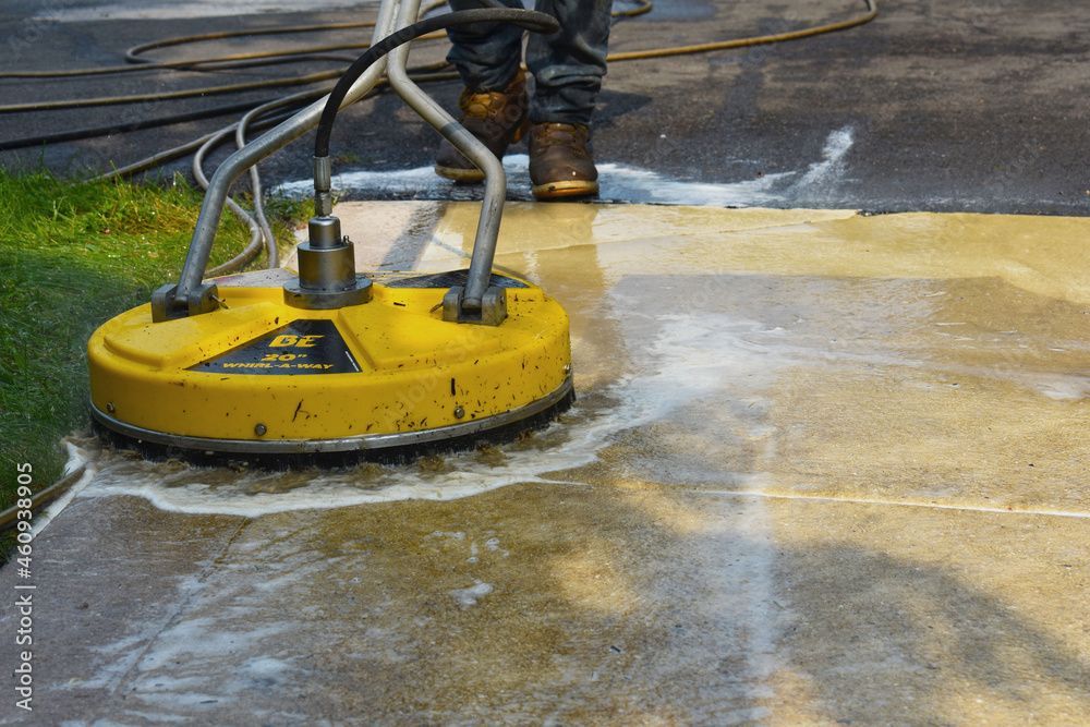 A worker uses a yellow circular pressure washer tool to clean a dirty concrete surface outdoors.