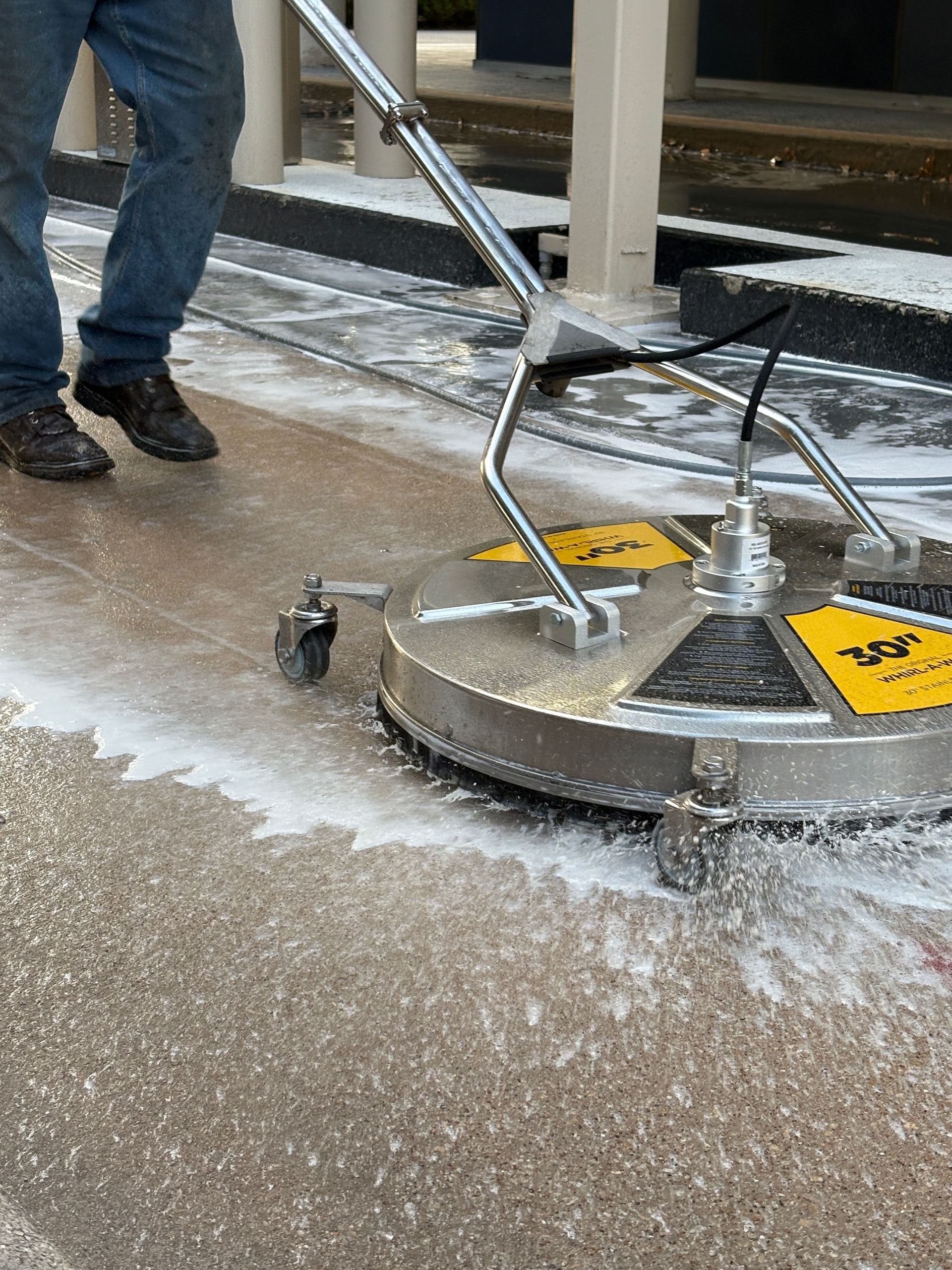 Person pushing a metal floor scrubber over a wet concrete surface indoors