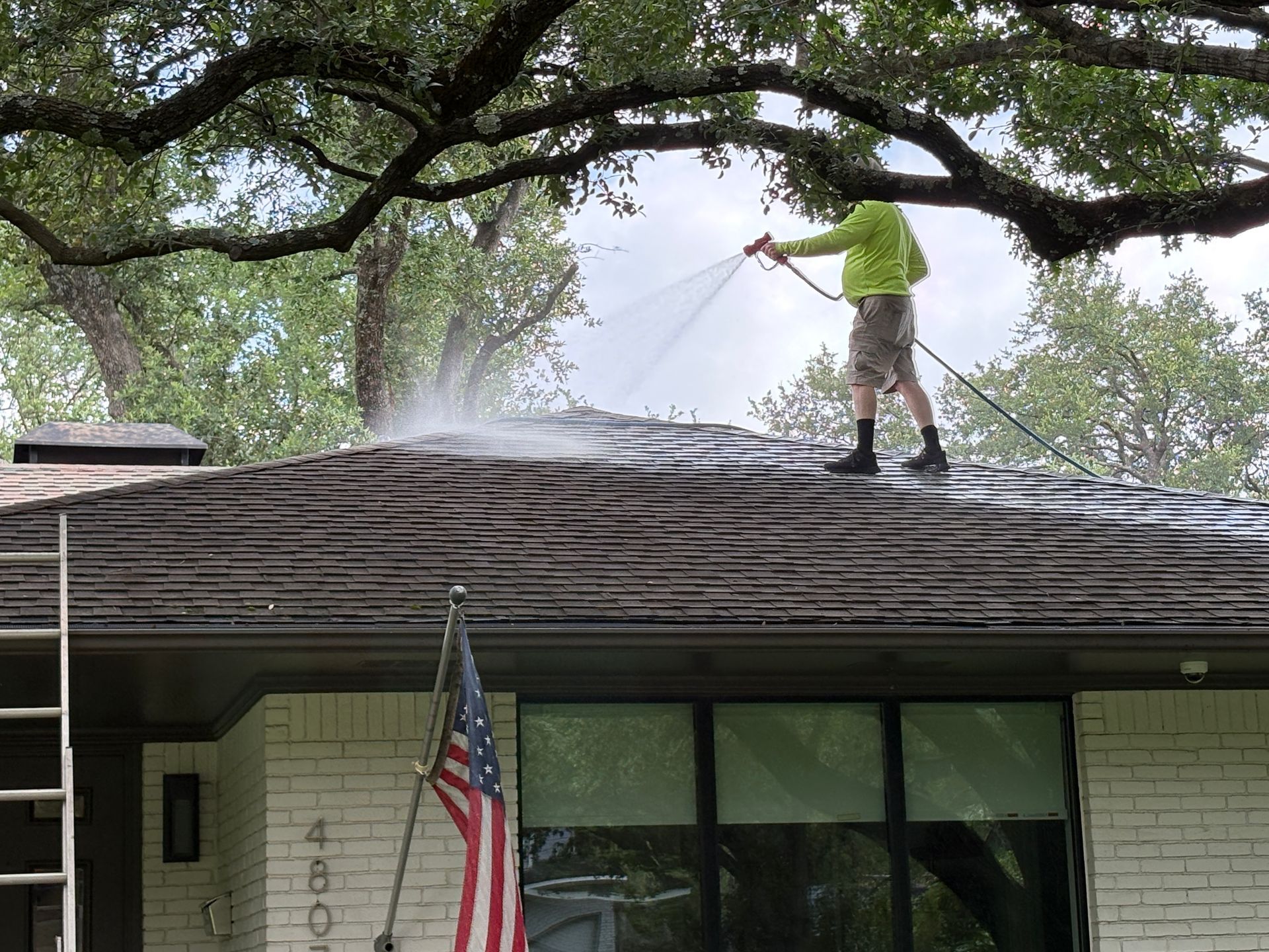 Worker pressure-washing a house roof under tree branches while an American flag hangs below