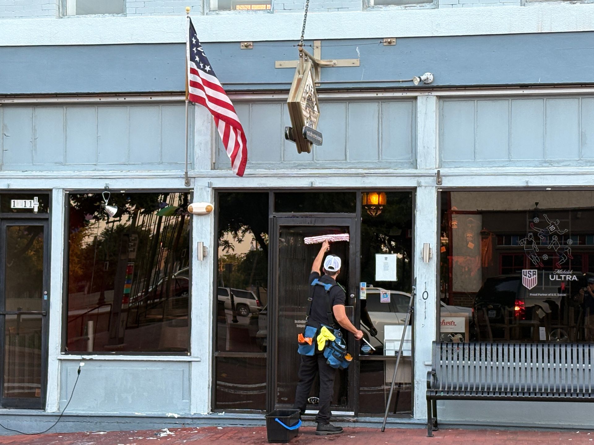 Person standing in a storefront doorway beneath an American flag and a hanging sign