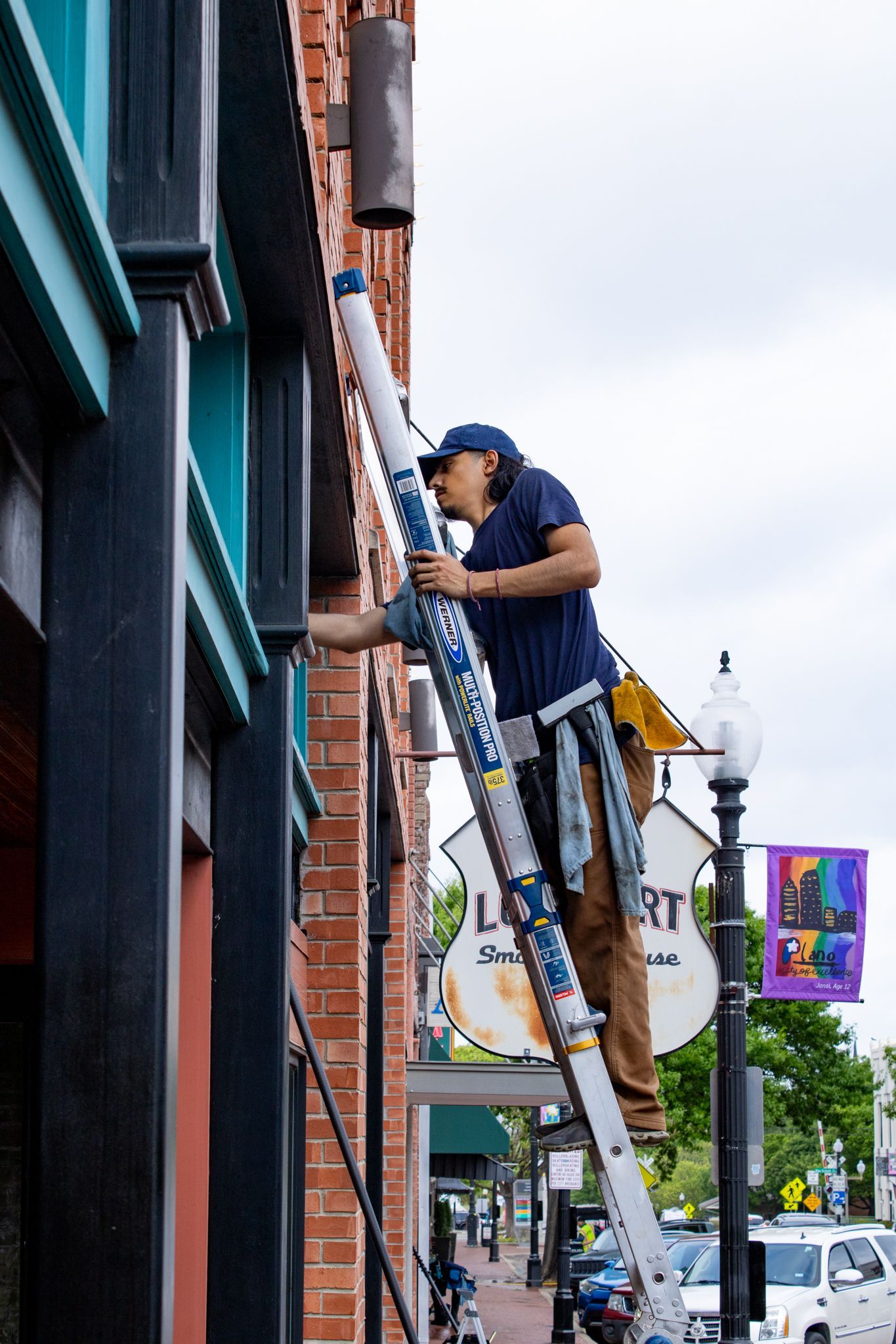 Worker on a tall ladder painting a brick building exterior on a city street.