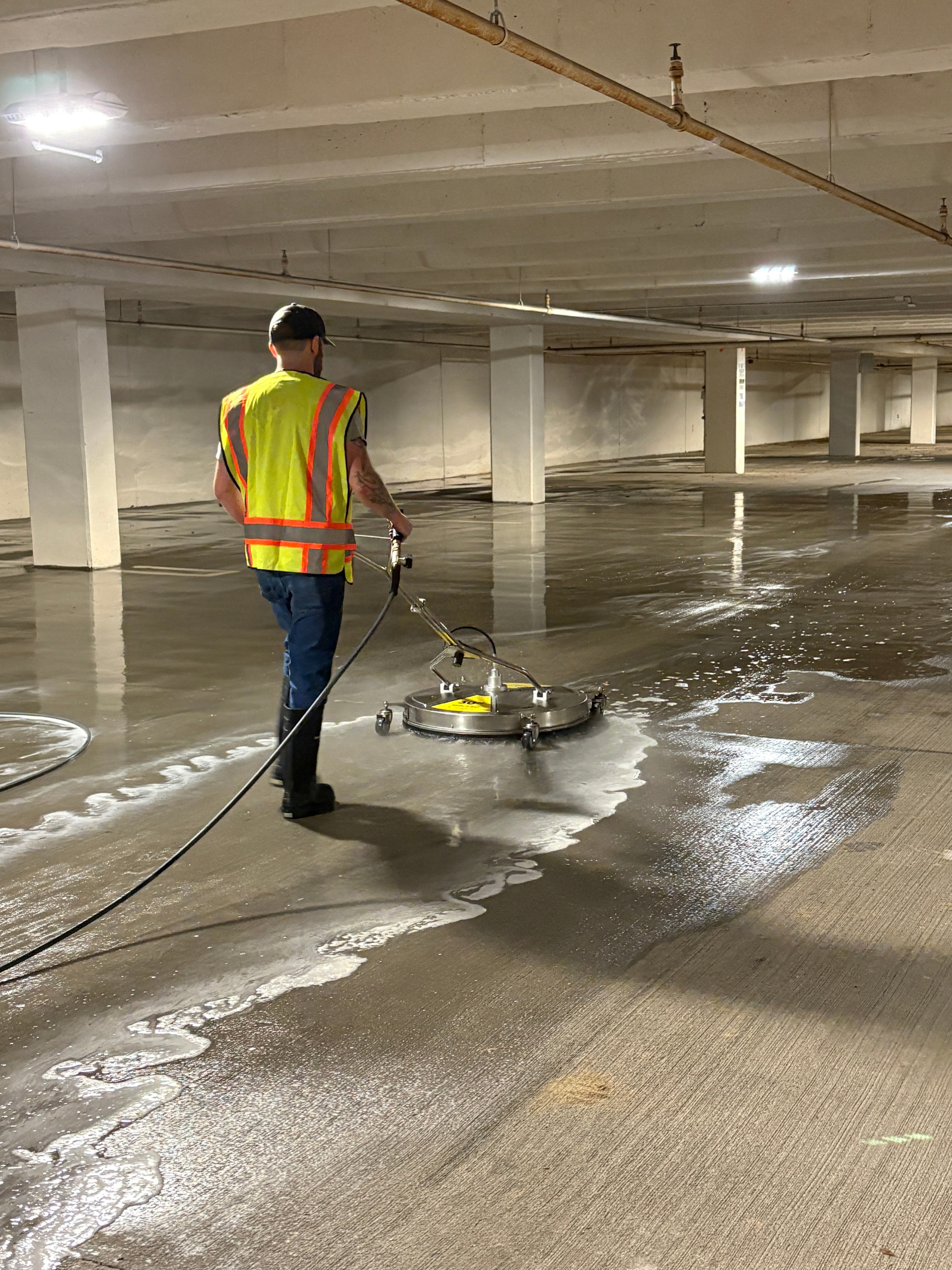 Worker in a reflective vest operating a floor scrubbing machine in a wet parking garage.