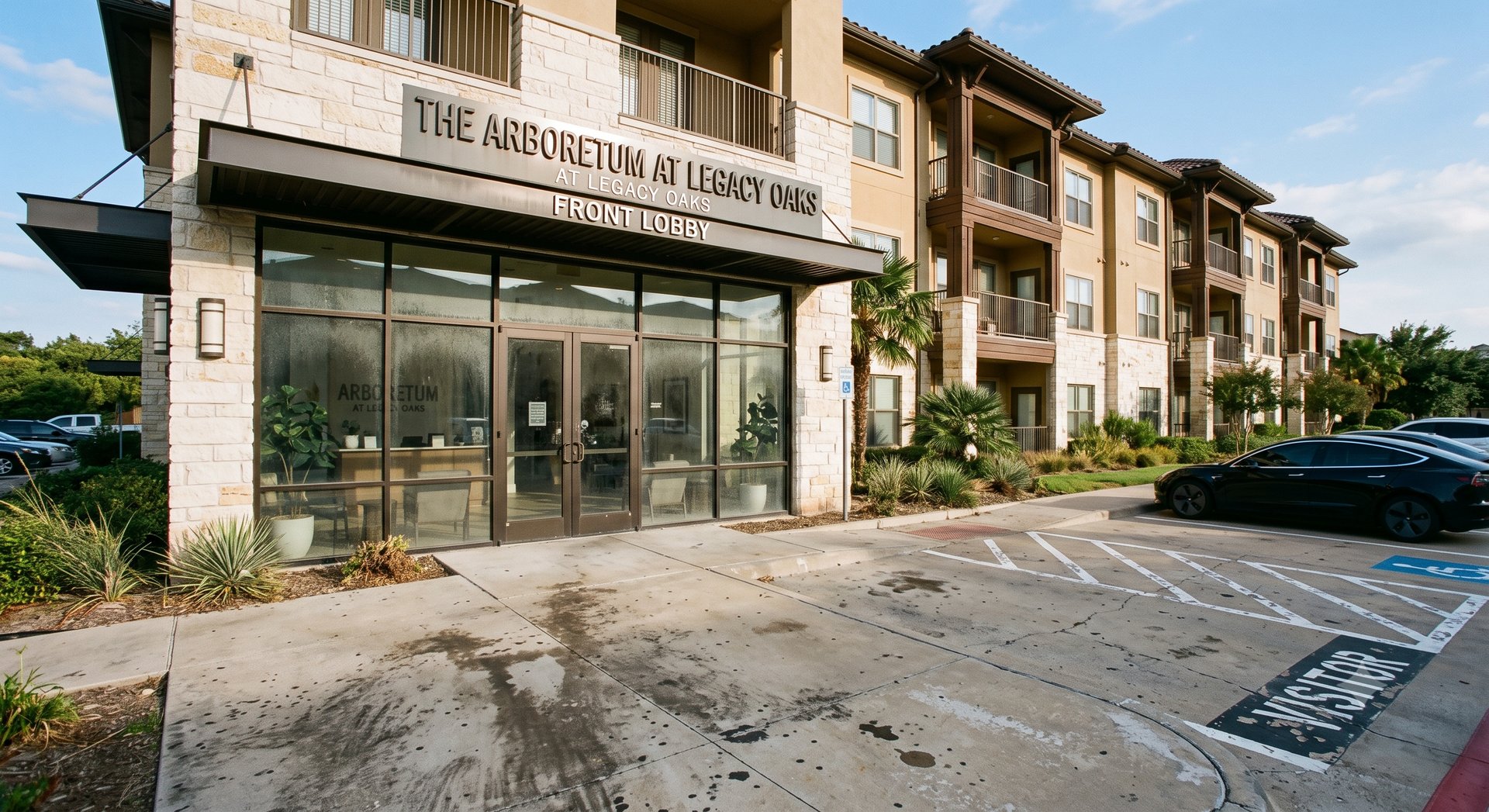 Apartment complex entrance with glass doors, beige stucco facade, and parked cars in front