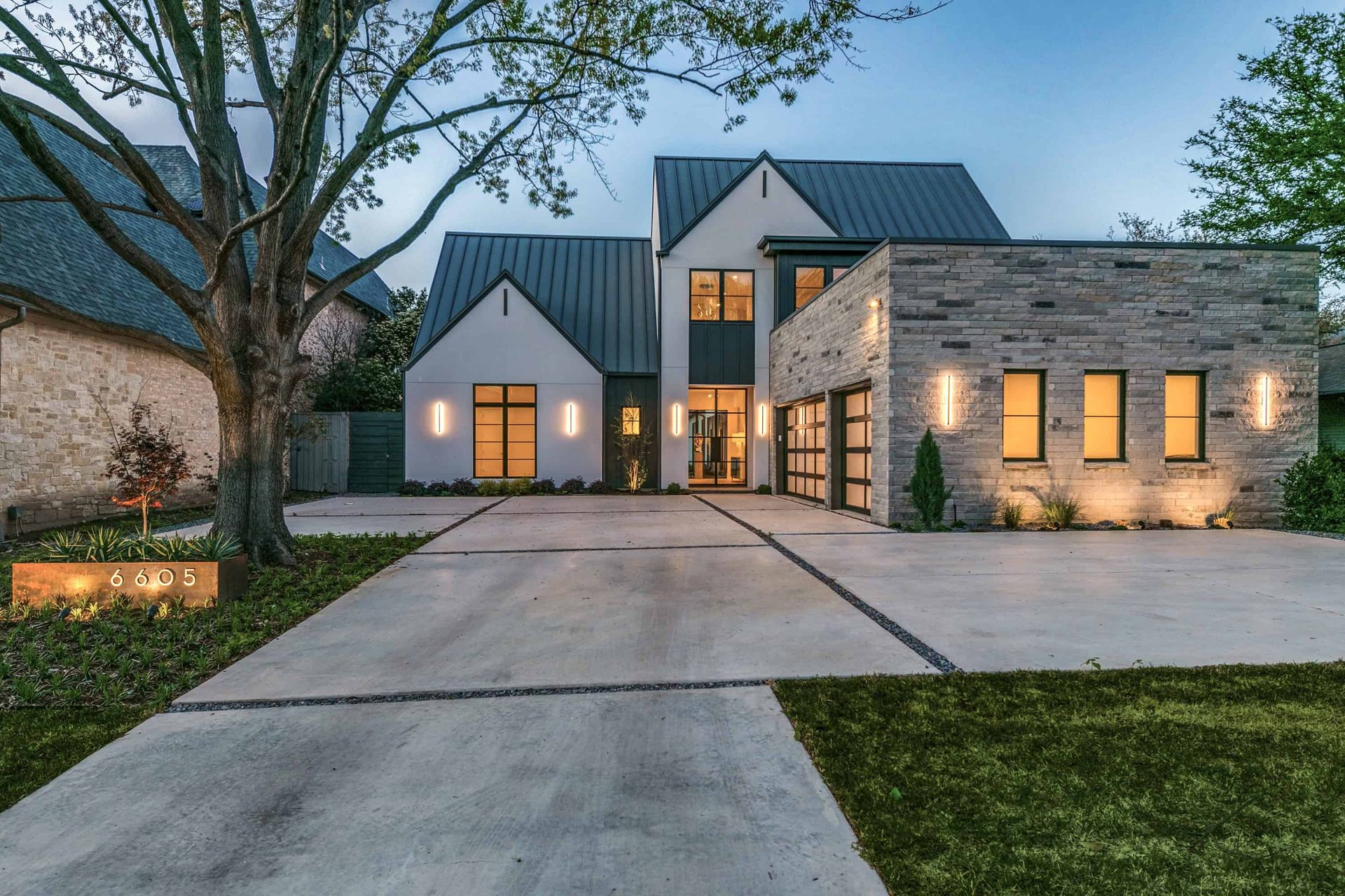 Modern stone-and-gray house at dusk with warm lit windows, driveway, and front lawn