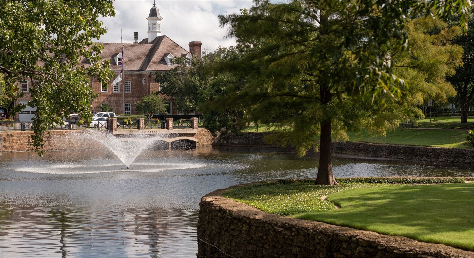 Park pond with a fountain, trees, and a brick building in the background.