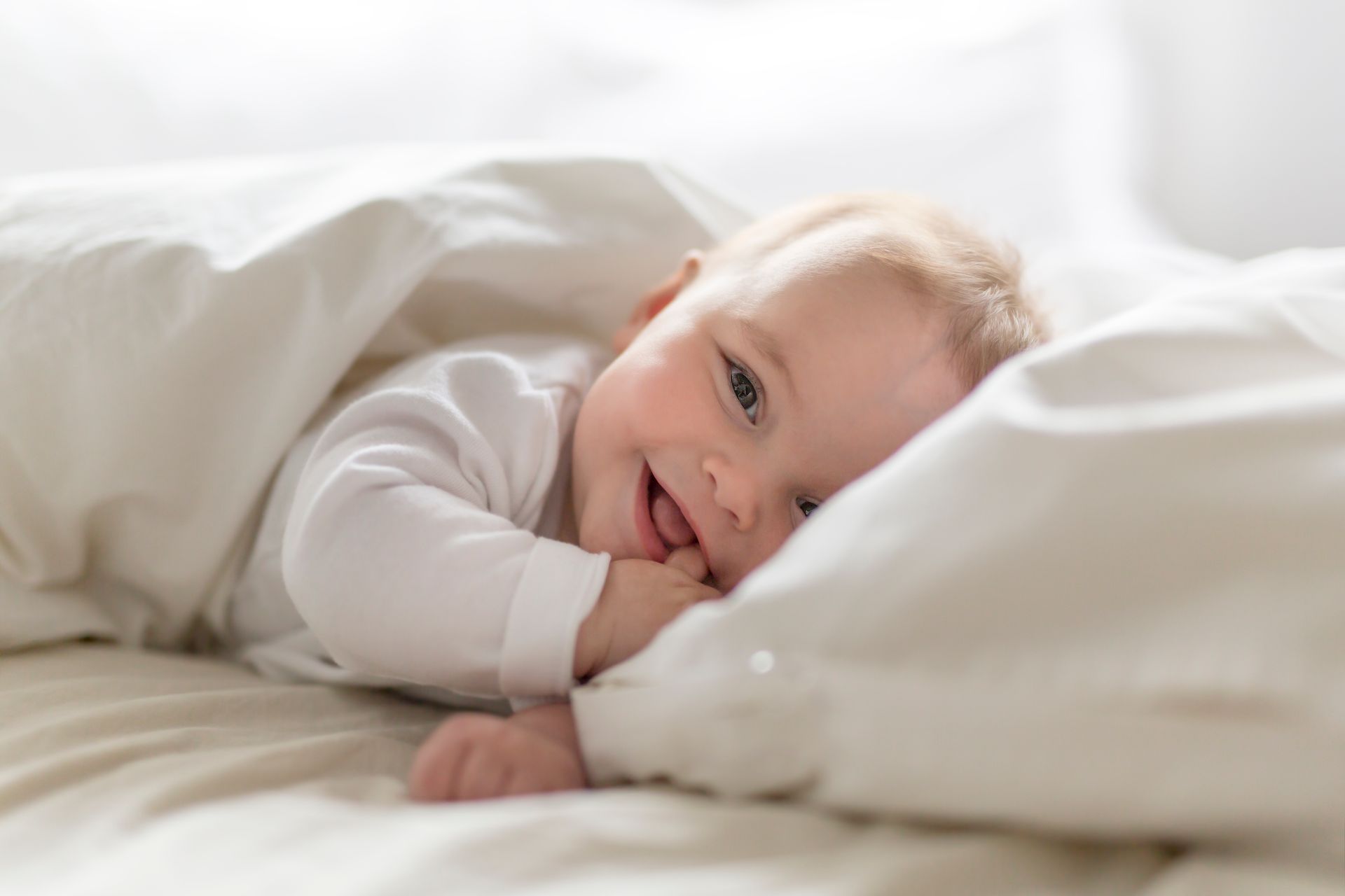 A smiling baby is lying under blankets