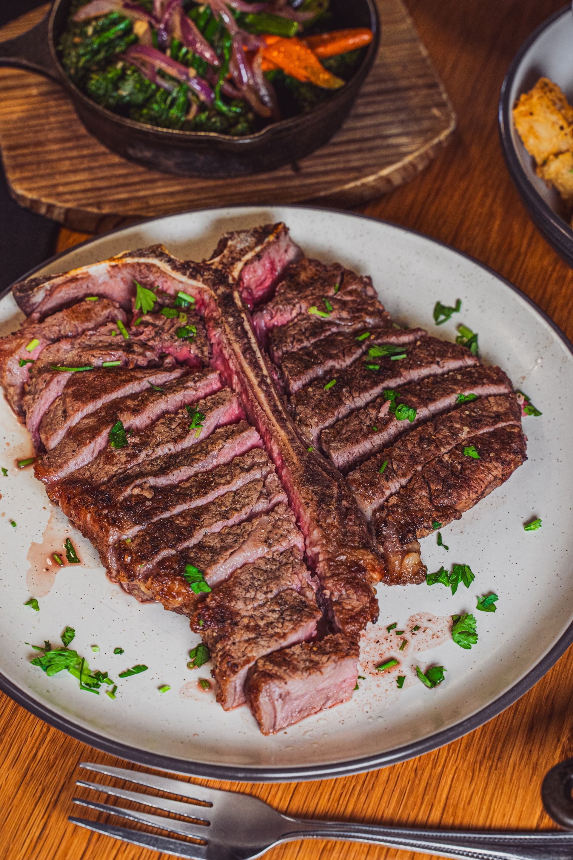 Grilled steak sliced on a white plate with herbs, served on a wooden table with a skillet of vegetables.