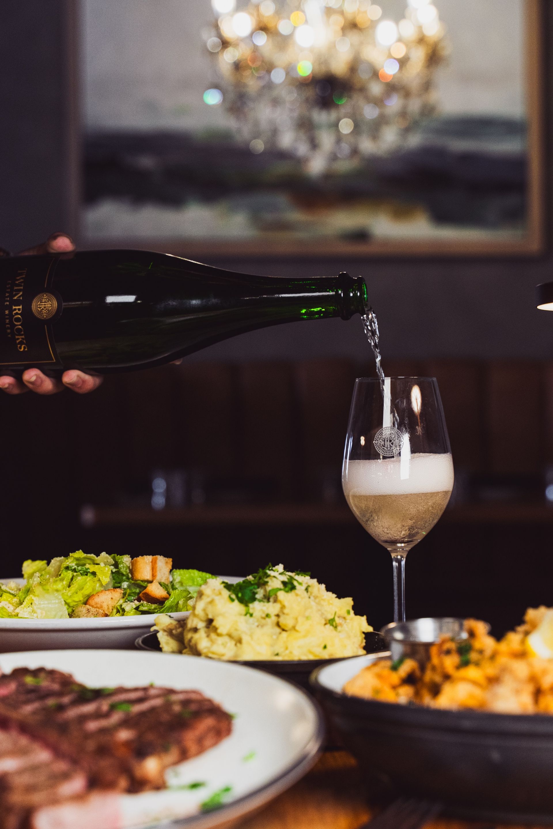 Wine being poured into a glass beside plated dinner dishes on a restaurant table