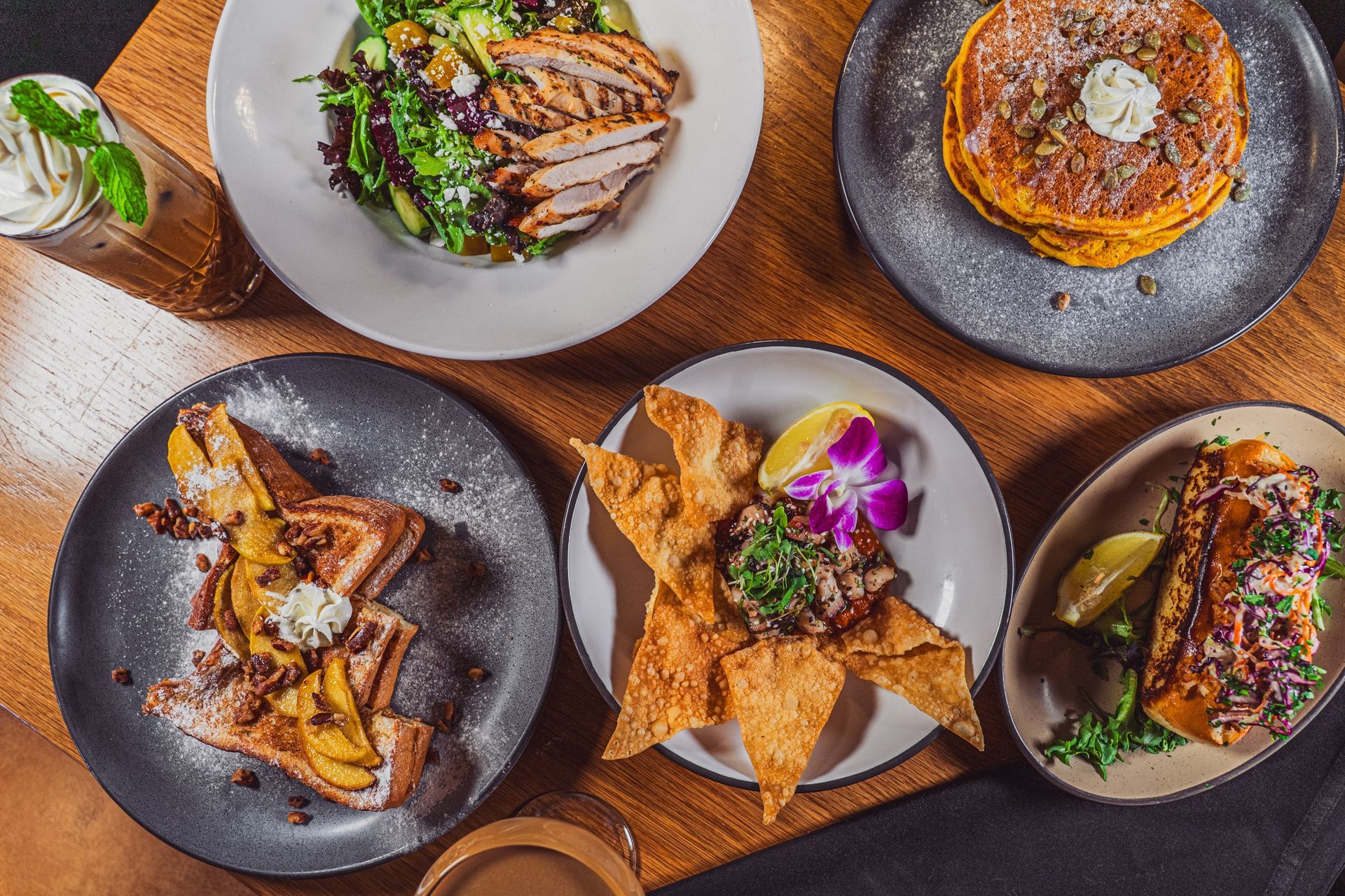 Assorted gourmet dishes on dark plates, including sandwiches, fries, and garnished entrées on a wooden table