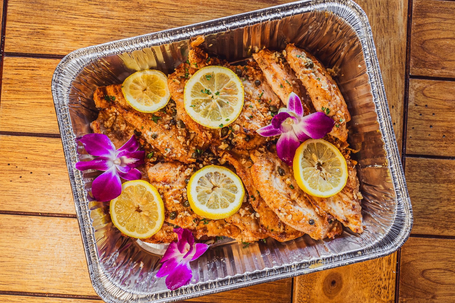 Chicken in a foil tray, topped with lemon slices and purple orchid flowers on a wooden table