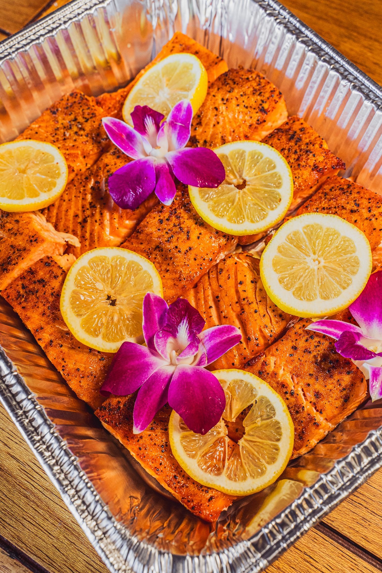 Baked fish fillets in a foil tray topped with lemon slices and purple orchid flowers