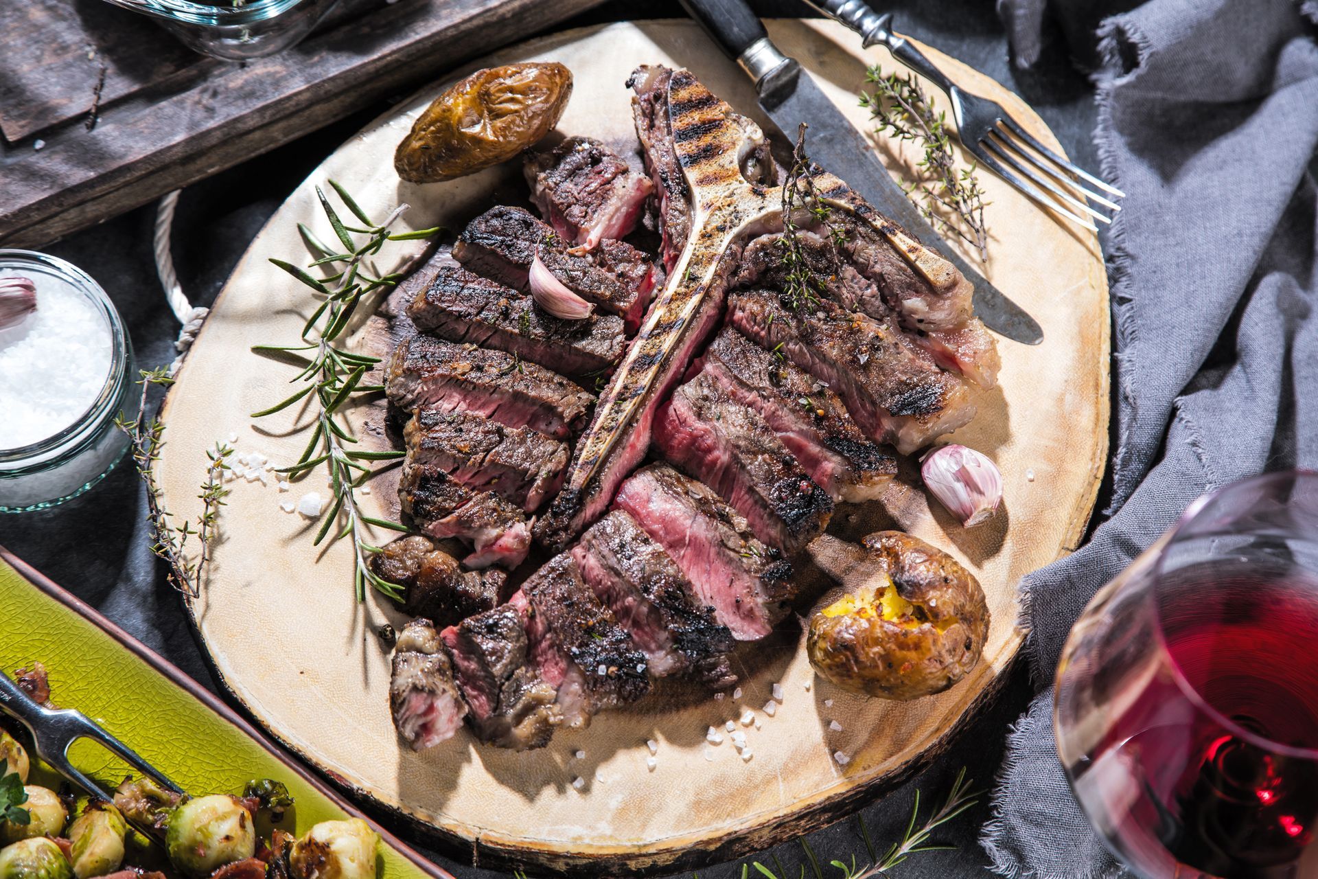 Grilled steak slices on a platter with rosemary, roasted potatoes, and a glass of red wine