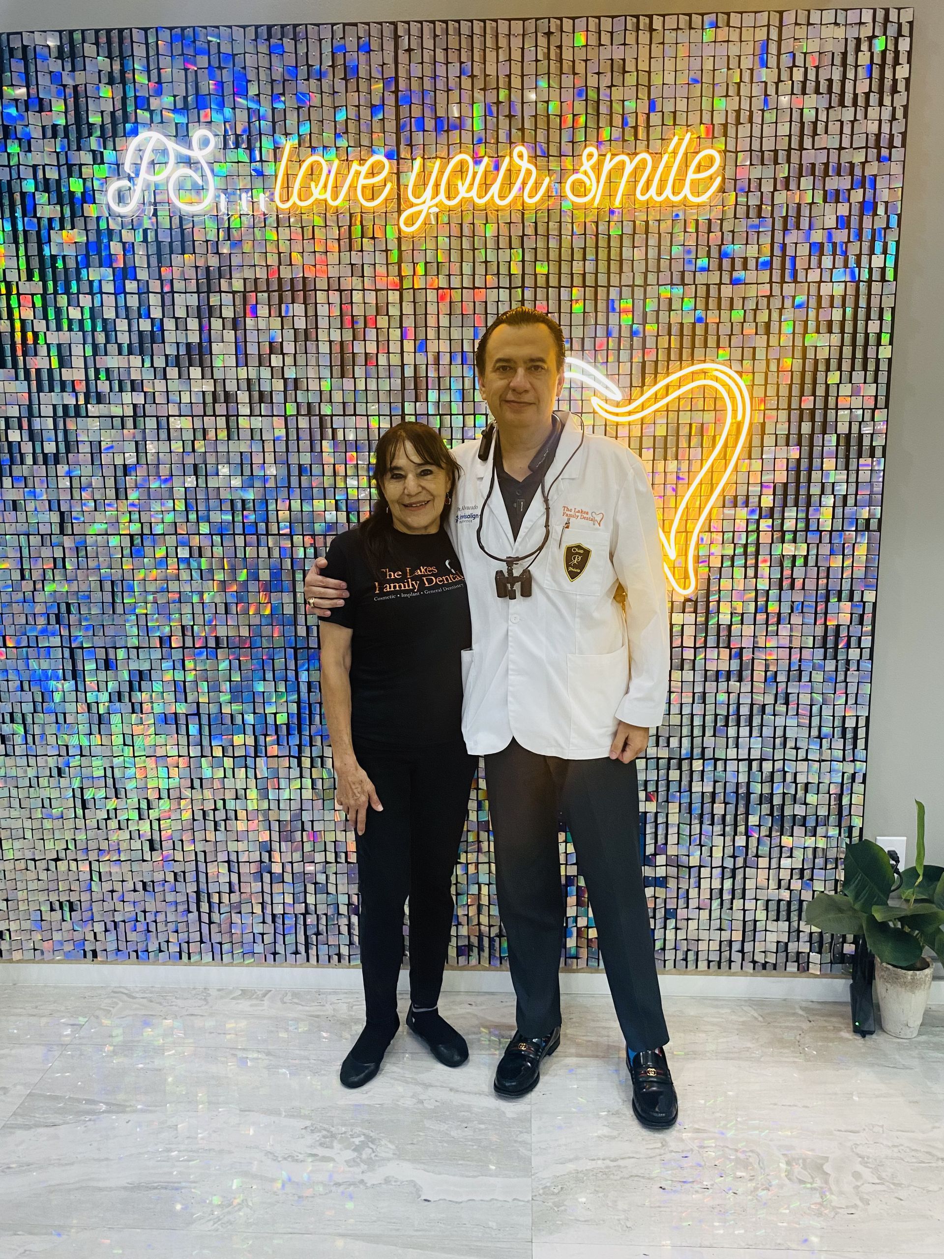 Two people smiling in front of a shimmering wall with neon sign that reads 