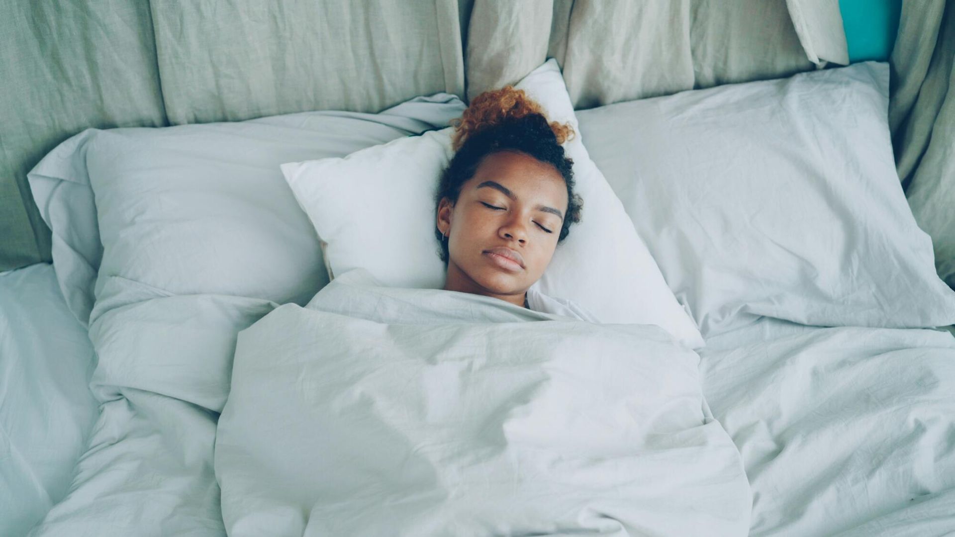 Person sleeping in bed, covered by a white blanket and surrounded by pillows.