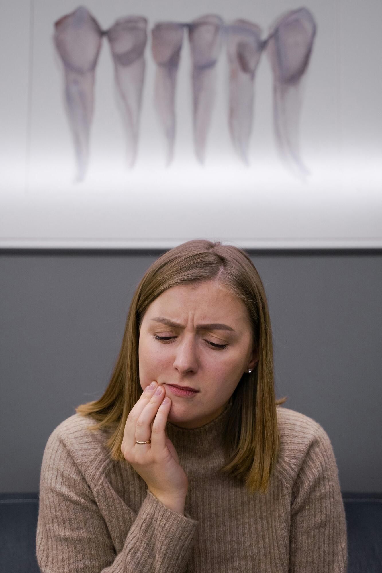 Woman holding her jaw in pain at dentist's office, teeth x-ray in background.
