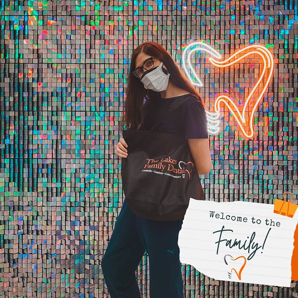 A woman wearing a mask and glasses is standing in front of a wall with a sign that says welcome to the family.