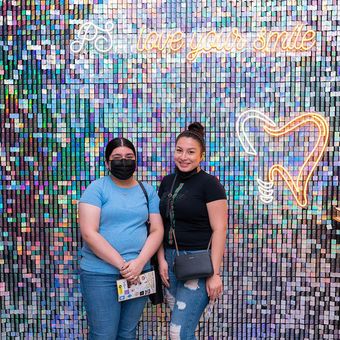 Two women are standing in front of a wall with a neon sign that says `` love your smile ''.