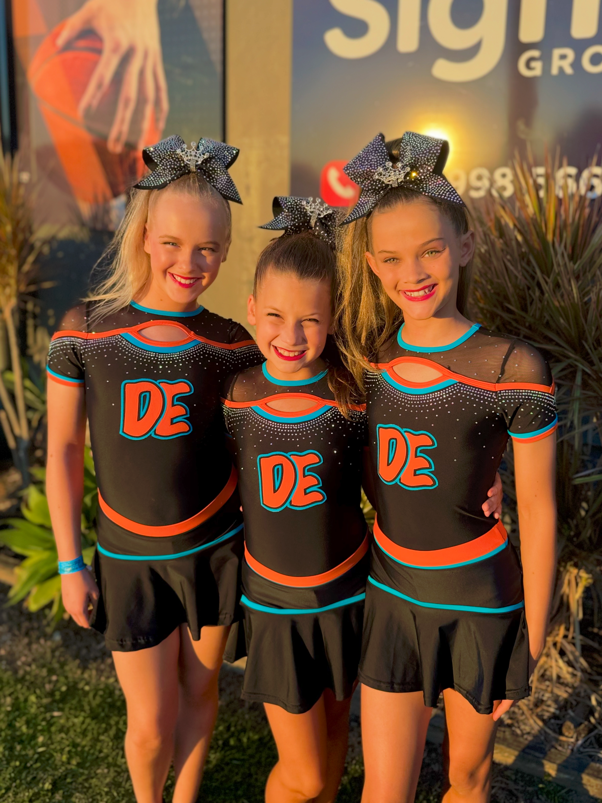 Three Young Cheerleaders In Matching Black And Orange Uniforms, Smiling, Outdoors — Dance Express Mackay In North Mackay, QLD