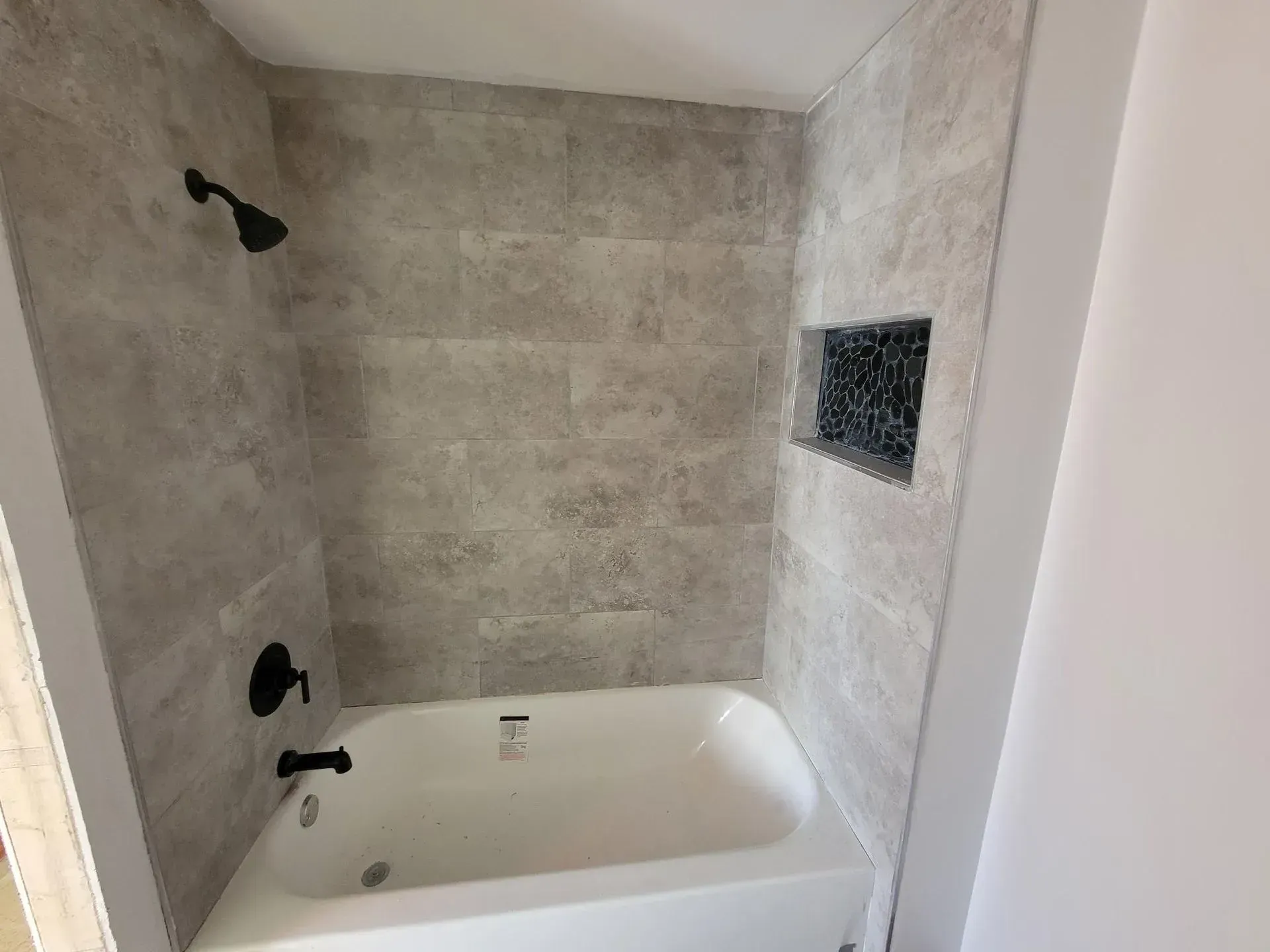 Bathroom shower with gray tiled walls, black fixtures, and a white tub.
