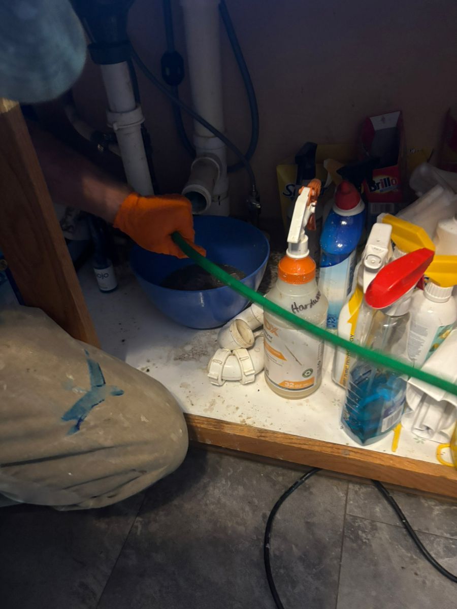 Plumber working under a sink with orange gloves and green tubing among cleaning supplies