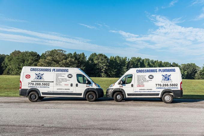 Two white plumbing service vans parked on grass, blue sky background. Vans have company logo and phone number on the side.