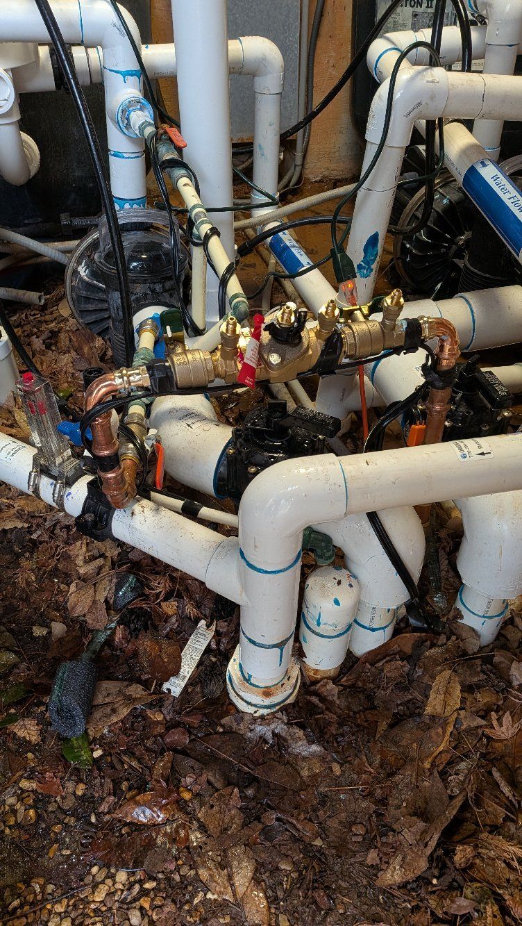 Close-up of outdoor plumbing pipes and valves, with white PVC and brass fittings amid wet leaves and dirt.