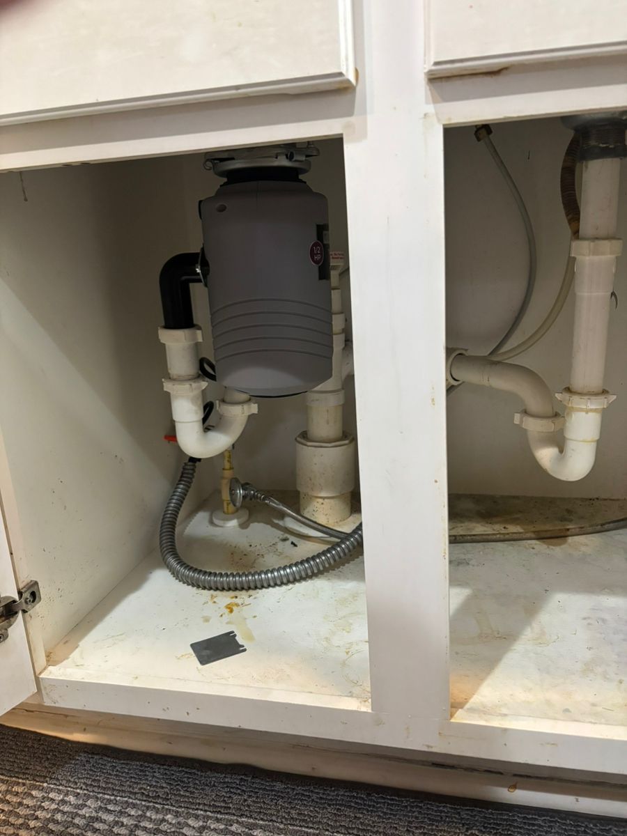 Under-sink plumbing with drain pipes, hoses, and a gray garbage disposal unit in a white cabinet.