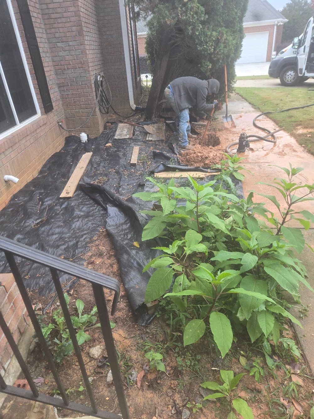 Person working in a front yard beside a brick house, handling landscaping debris and mulch.