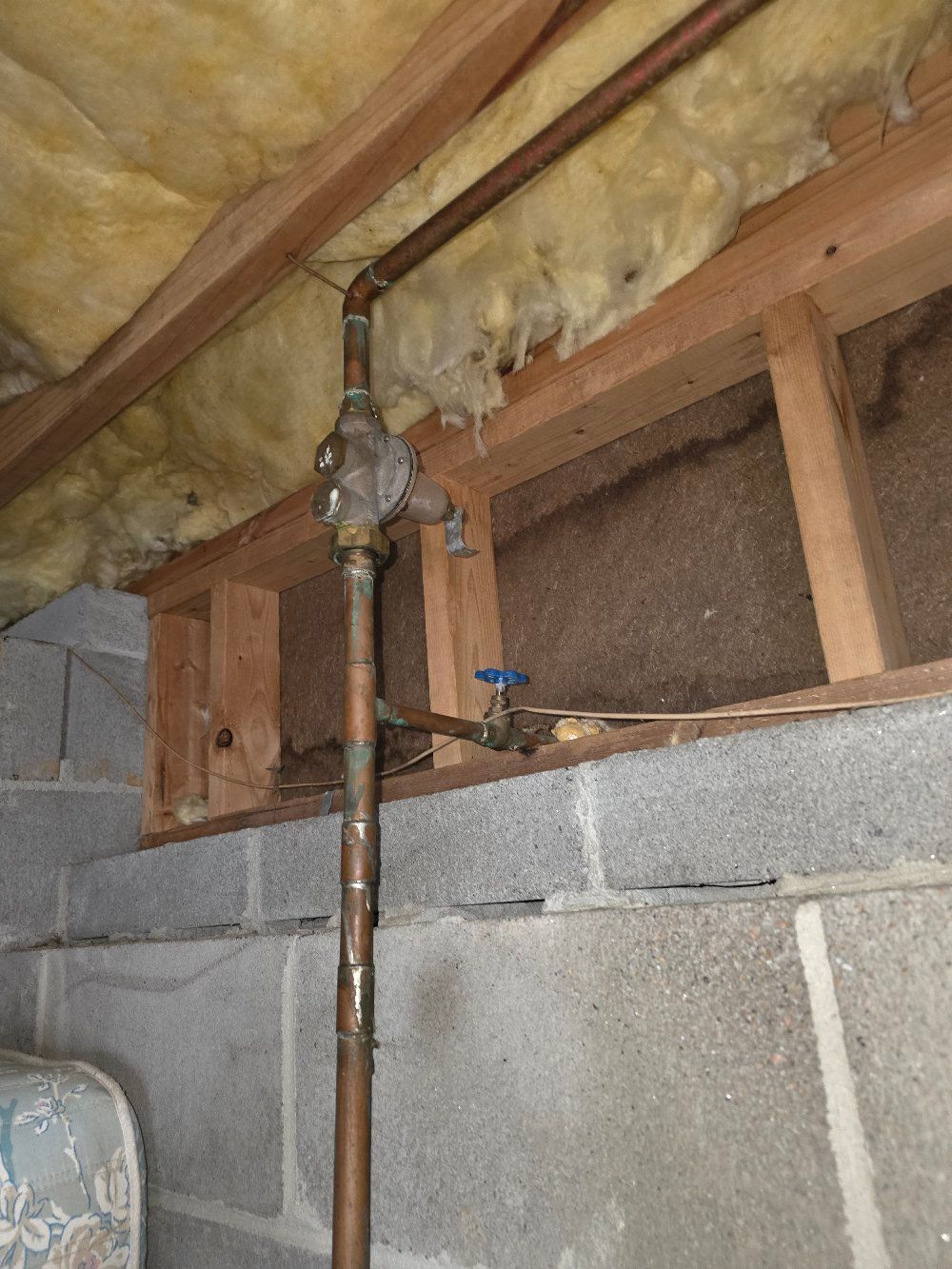 Basement wall with exposed copper pipes, a valve, cinder blocks, and insulation under rafters.