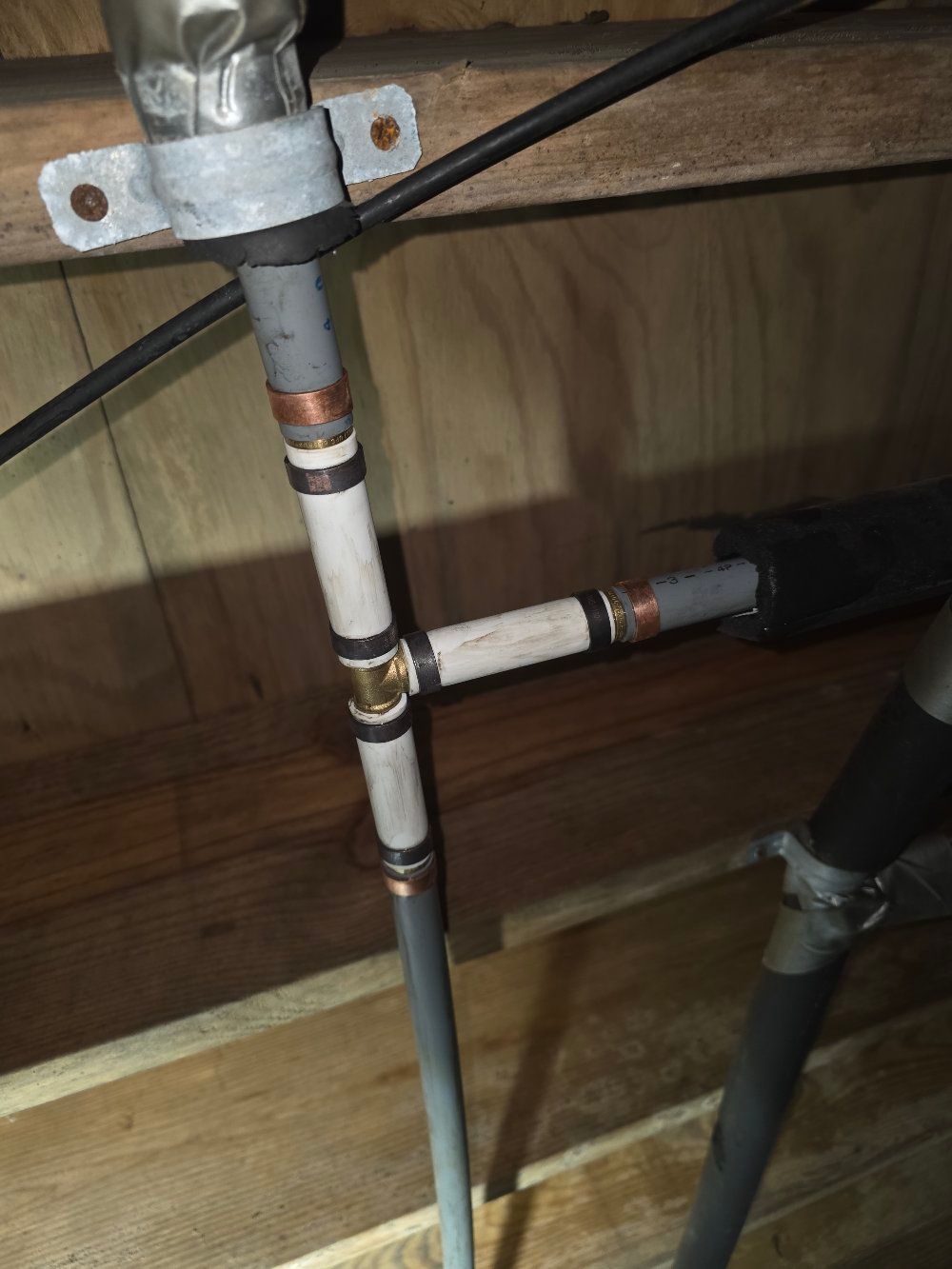 Close-up of a white PVC pipe fitting and valve assembly mounted under a wooden floor.
