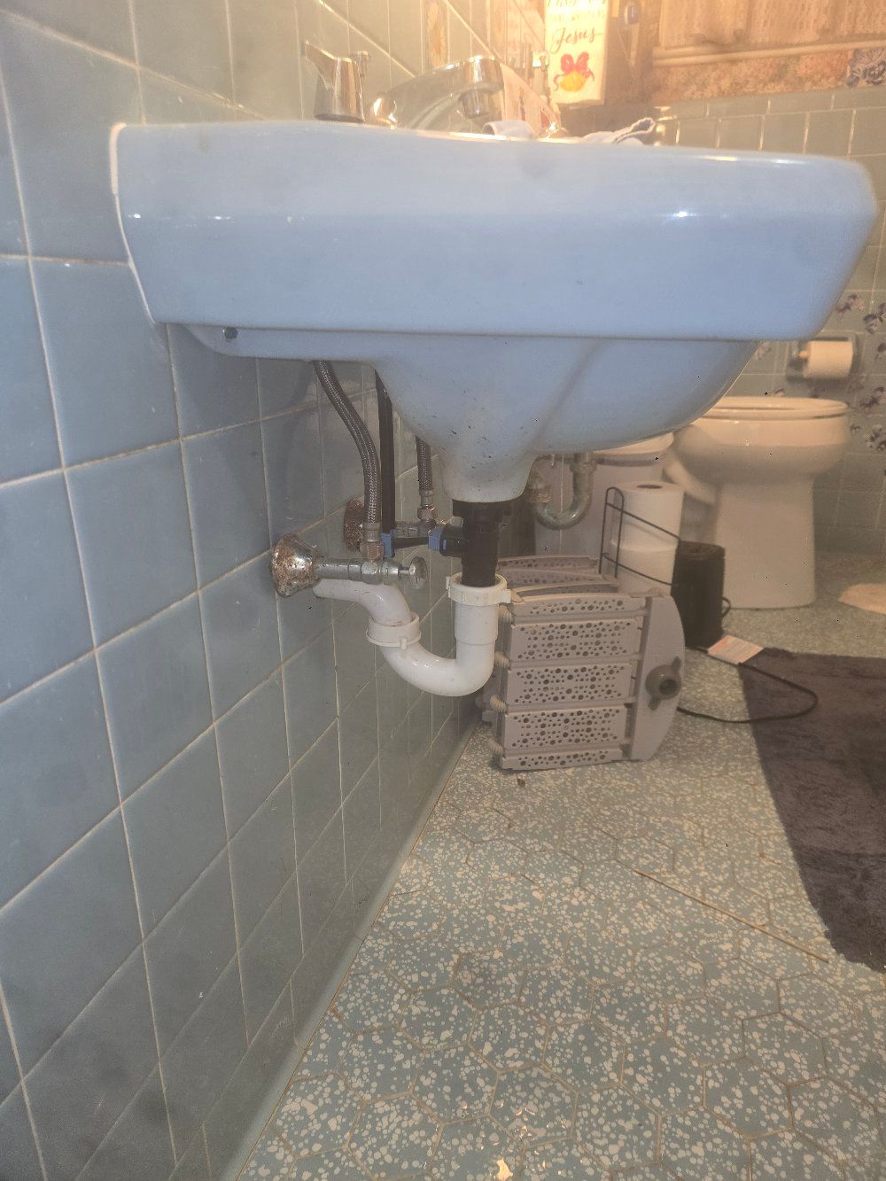 Blue wall-mounted sink in a tiled bathroom with exposed plumbing and a toilet in the background