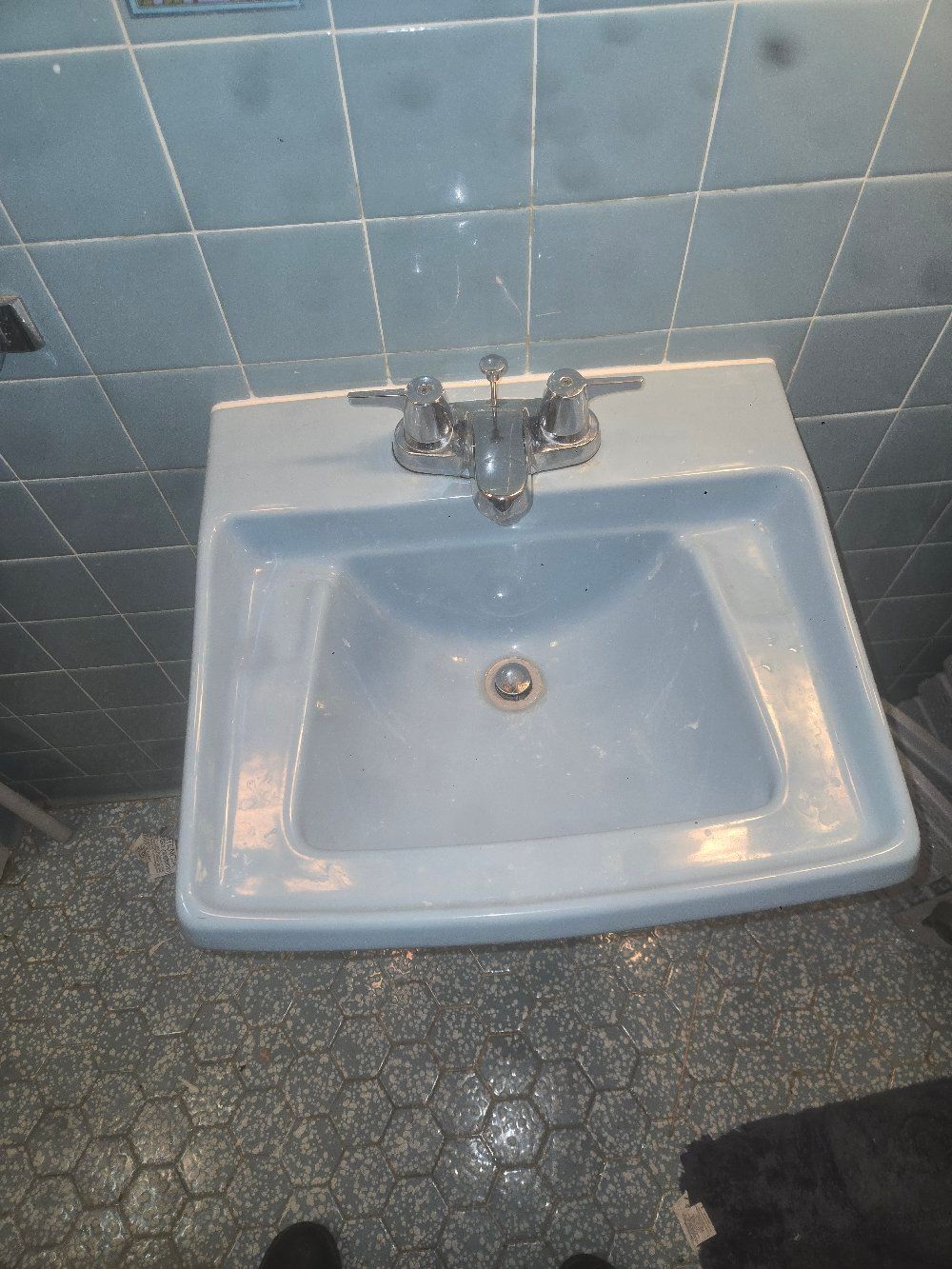 White porcelain bathroom sink with chrome faucet against blue tiled wall and dark speckled floor.