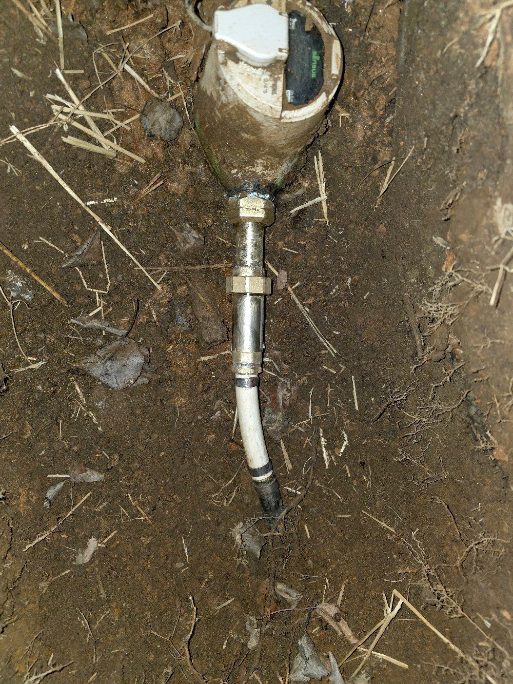 Close-up of a buried metal pipe fitting connected to a white cable in dirt.